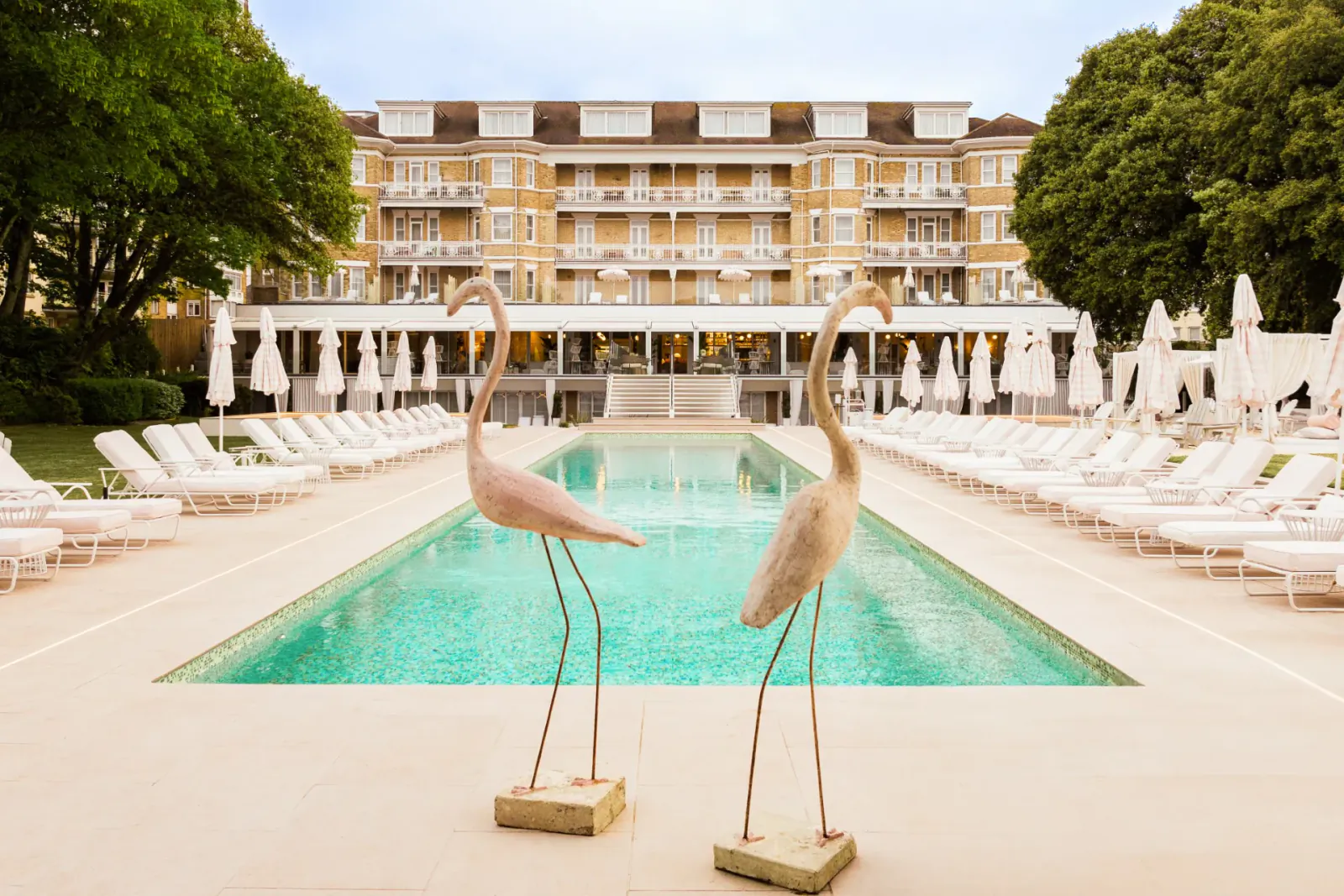 Two tall pink flamingo statues stand on either side of a long turquoise pool at a luxury hotel with lounge chairs and umbrellas