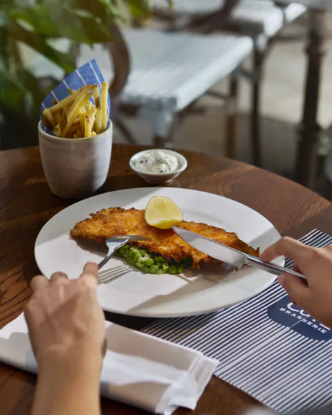 Hands cutting a breaded fish fillet with lemon and peas on a white plate at Côte Brasserie, with fries and tartar sauce on a wooden table