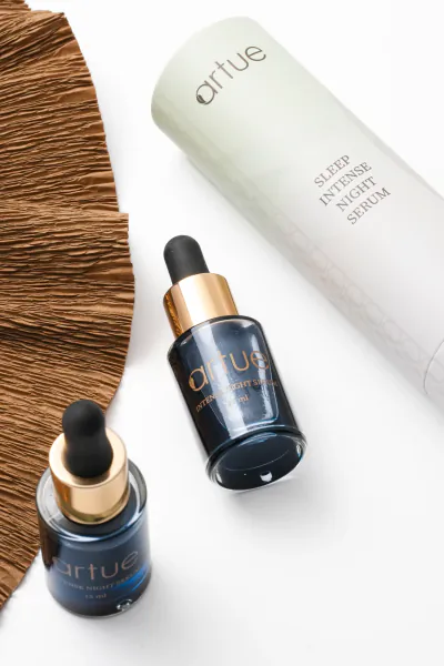 Artue skincare bottles: two dark blue Intense Night Serum dropper bottles and a light green Sleep Intense Night Serum tube beside a brown palm leaf on white background