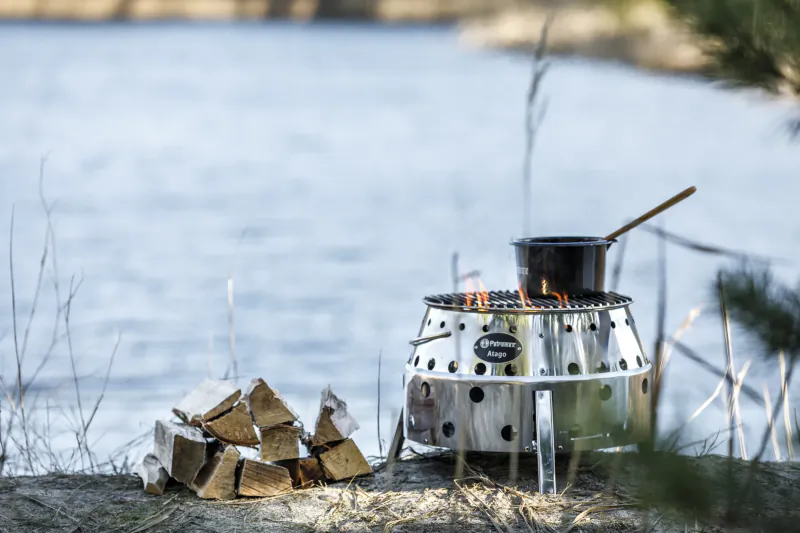 Portable stainless steel fire pit with burning logs and a black pot on top, stacked firewood beside a calm lake