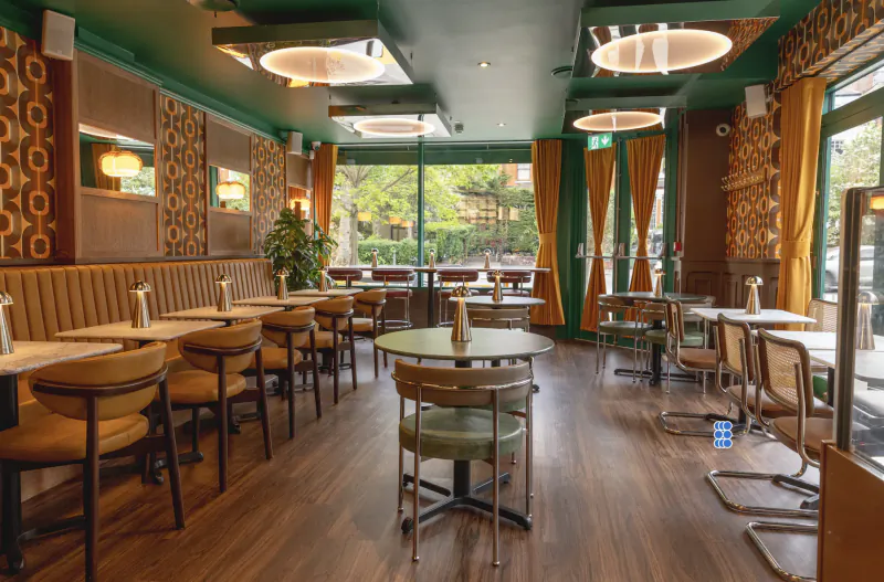 Modern Italian restaurant interior with green walls, patterned wallpaper, round tables, and large windows overlooking trees