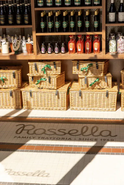 Rossella Family Trattoria storefront with wicker gift baskets and wine bottles on wooden shelves, logo on tiled floor