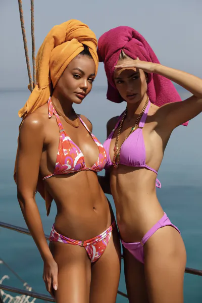 Two women in colorful bikinis and towel-wrapped heads pose confidently on a boat deck overlooking the sea