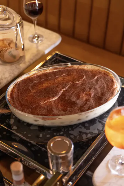 Large tiramisu in a glass dish dusted with cocoa, beside a glass of red wine and an orange cocktail on a marble table