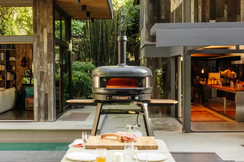 Gozney pizza oven with open flame on patio table, raw pizza dough and toppings on cutting board in foreground