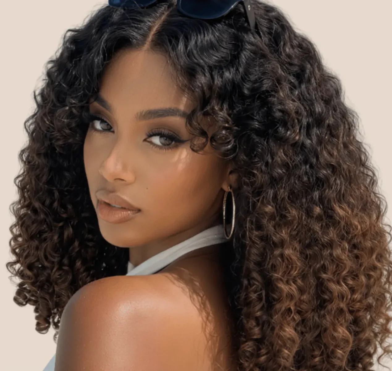 Woman with voluminous curly brown hair wearing sunglasses on her head and gold hoop earrings