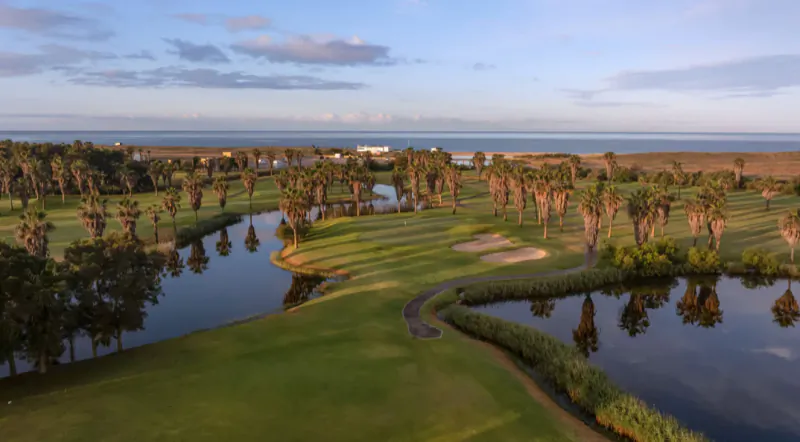 Aerial view of a lush golf course with palm trees, water hazards, and ocean view at sunset