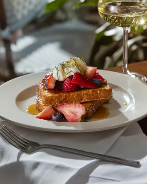 French toast topped with strawberries, blueberries, whipped cream and syrup on a white plate with a glass of white wine