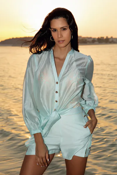 Woman in light blue satin shirt and shorts posing confidently by the water at golden hour sunset