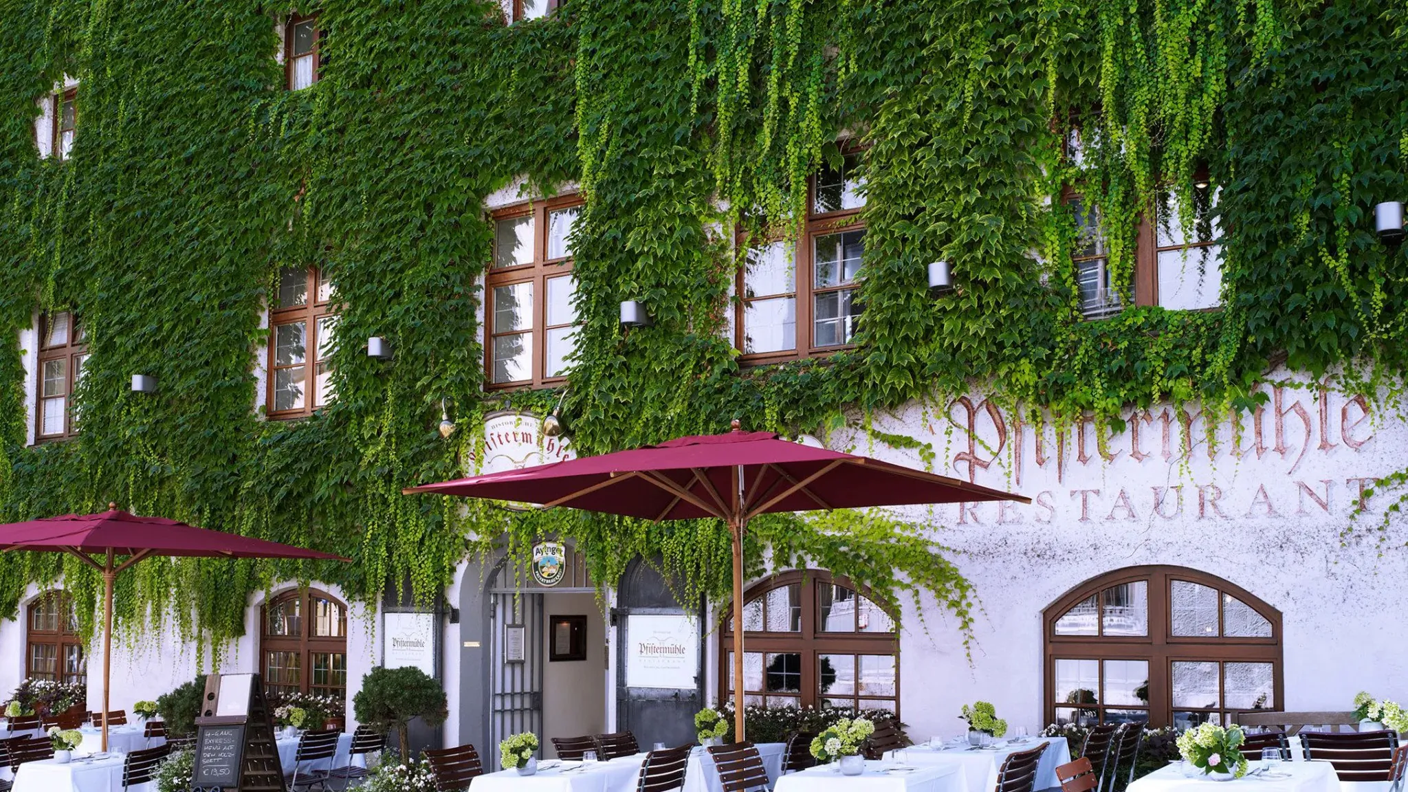 Platzl Hotel Munich exterior with ivy-covered facade, red umbrellas, and outdoor restaurant seating