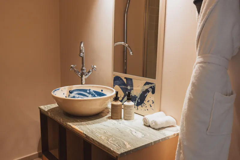 Modern hotel bathroom with a blue and white ceramic vessel sink on a marble vanity, chrome faucet, toiletries, folded towels, and a white bathrobe beside a mirror with blue abstract art