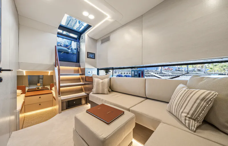 Modern yacht interior with L-shaped beige sofa, striped pillows, wooden coffee table, illuminated staircase, and large windows overlooking a marina