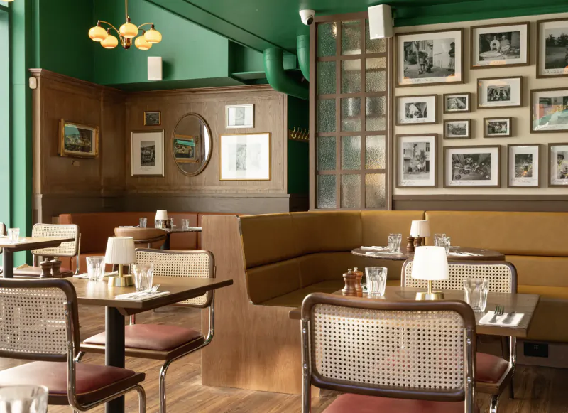 Interior of a stylish Italian restaurant with green walls, wooden booths, cane chairs, framed photos, and set tables