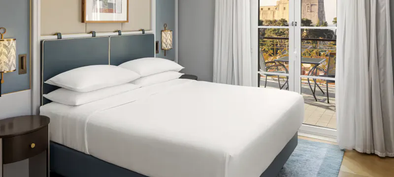 Hotel bedroom with large white bed, blue headboard, and balcony overlooking historic buildings