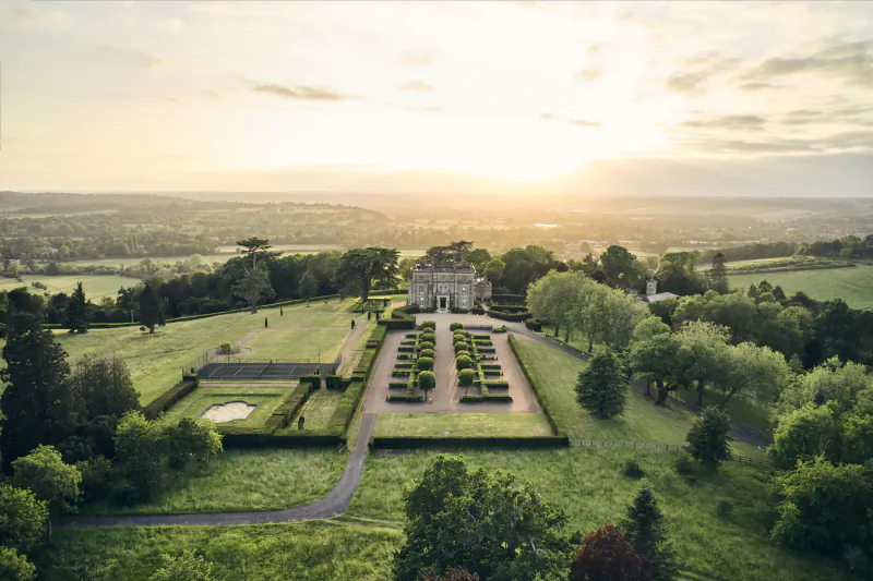 Aerial view of Hedsor House at sunset, grand estate with formal gardens, geometric hedges, and rolling countryside beyond