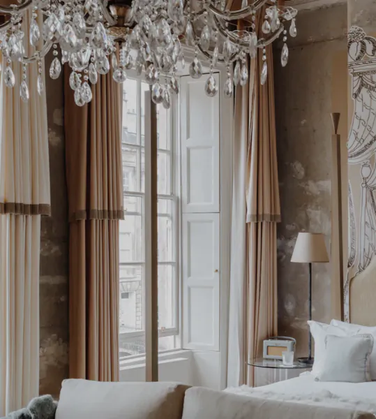 Luxurious hotel bedroom with crystal chandelier, tall windows with beige curtains, and a bed with white pillows
