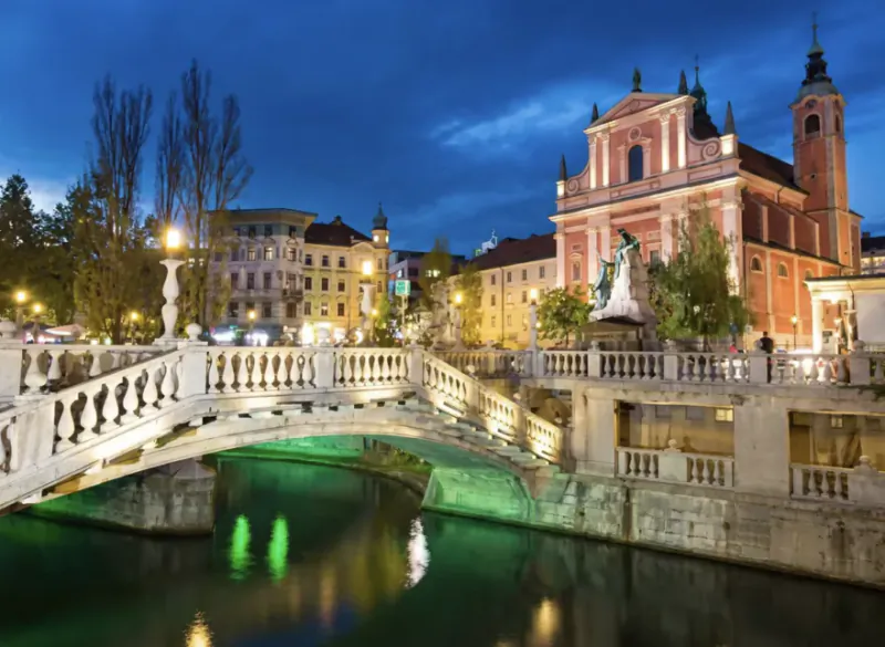 Illuminated stone bridge over a glowing green river at dusk, with a grand red church and statue in the background in Ljubljana, Slovenia