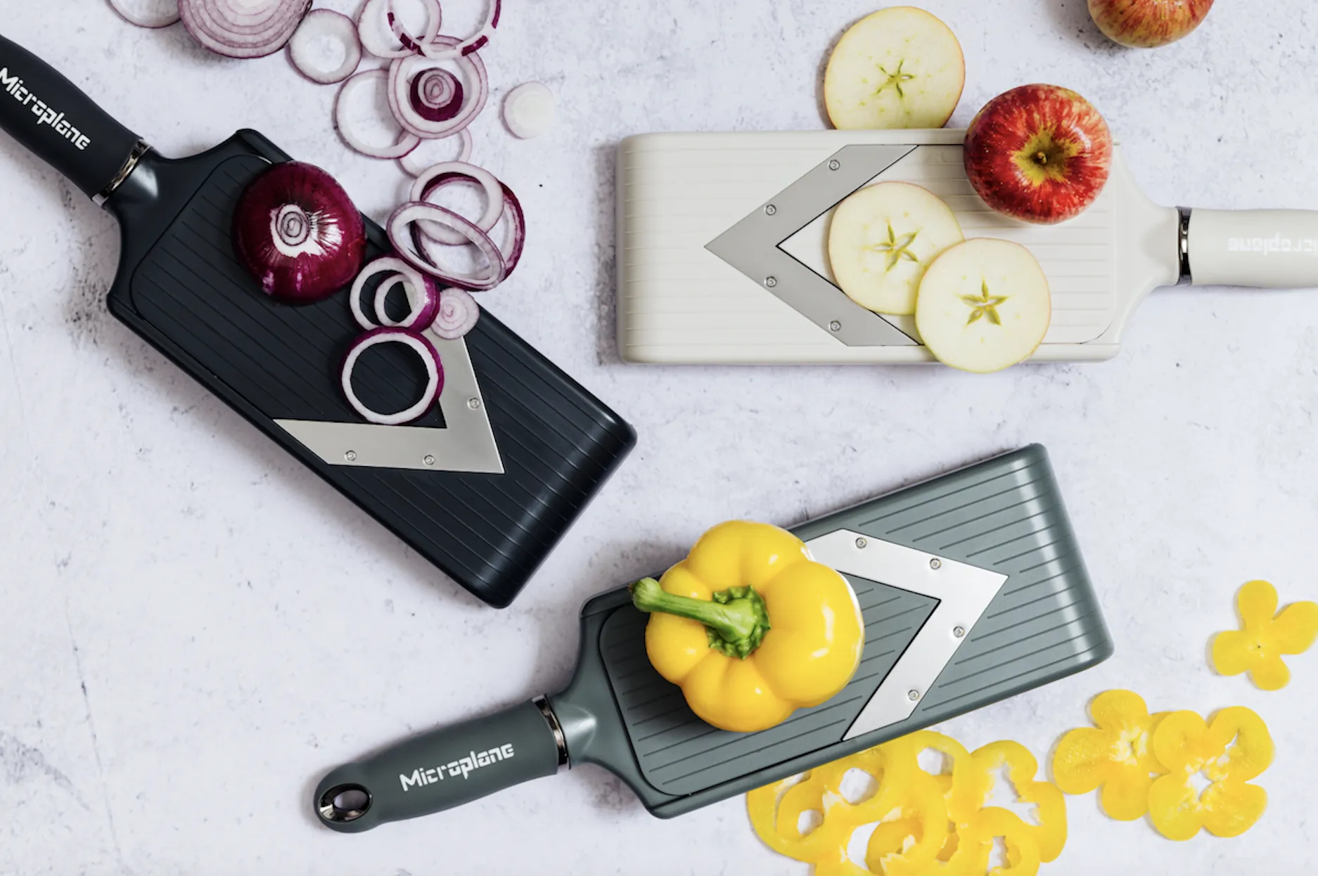 Three Microplane V-Blade slicers with sliced red onion, apple, and yellow bell pepper on marble