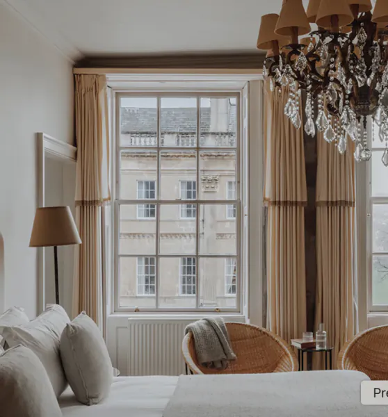 Elegant hotel bedroom with large window overlooking historic Bath buildings, beige curtains, crystal chandelier, wicker chairs and white bed linens