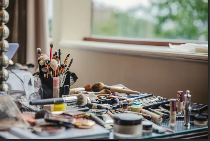 Makeup brushes, eyeshadow palettes, lipsticks and beauty products scattered on a vanity table near a window