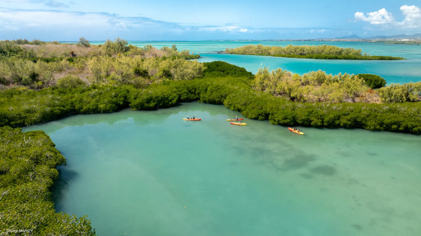 Aerial view of three kayakers paddling through turquoise waters surrounded by lush green mangroves and distant islands