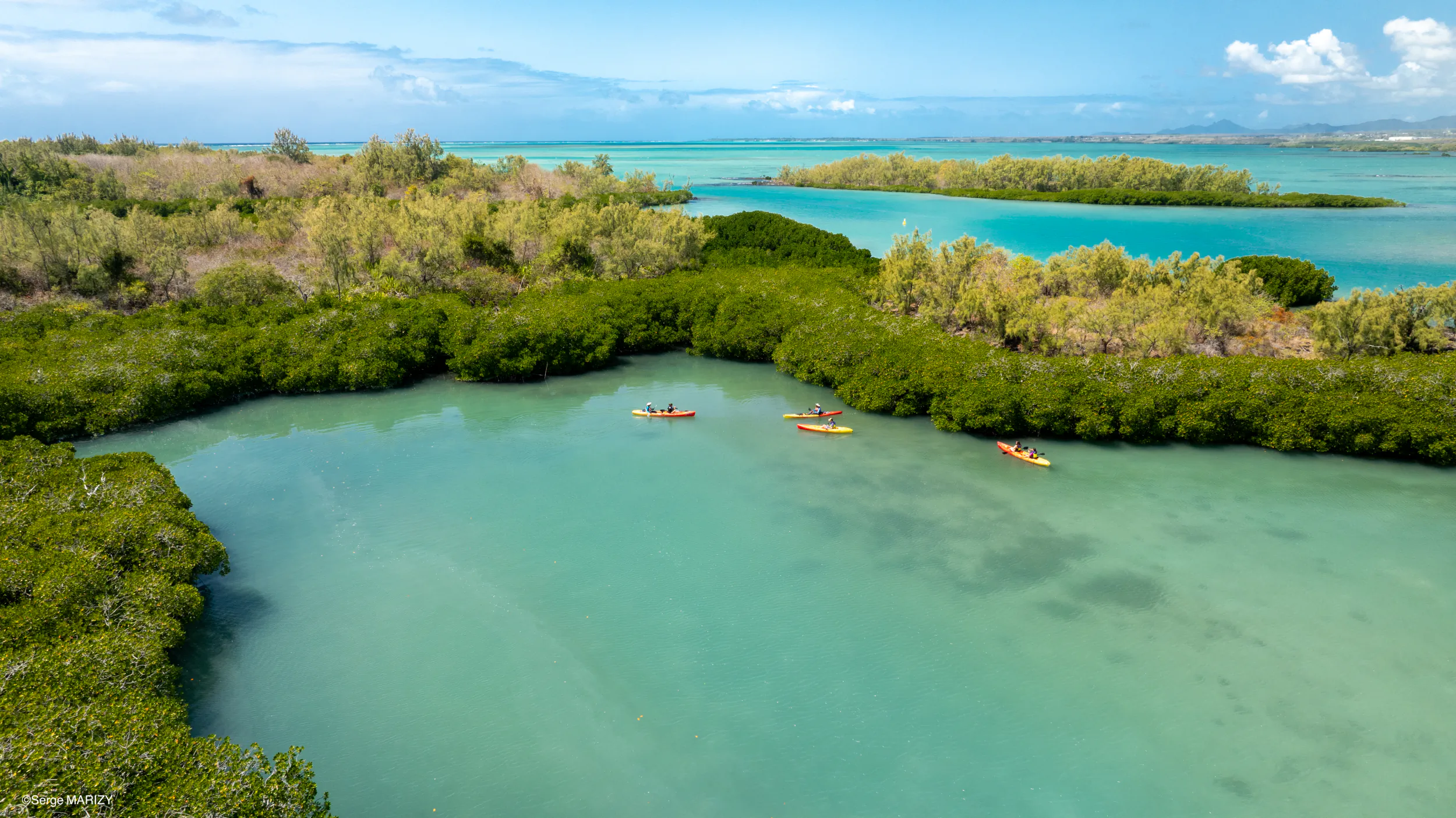 Aerial view of three kayakers paddling through turquoise waters surrounded by lush green mangroves and distant islands