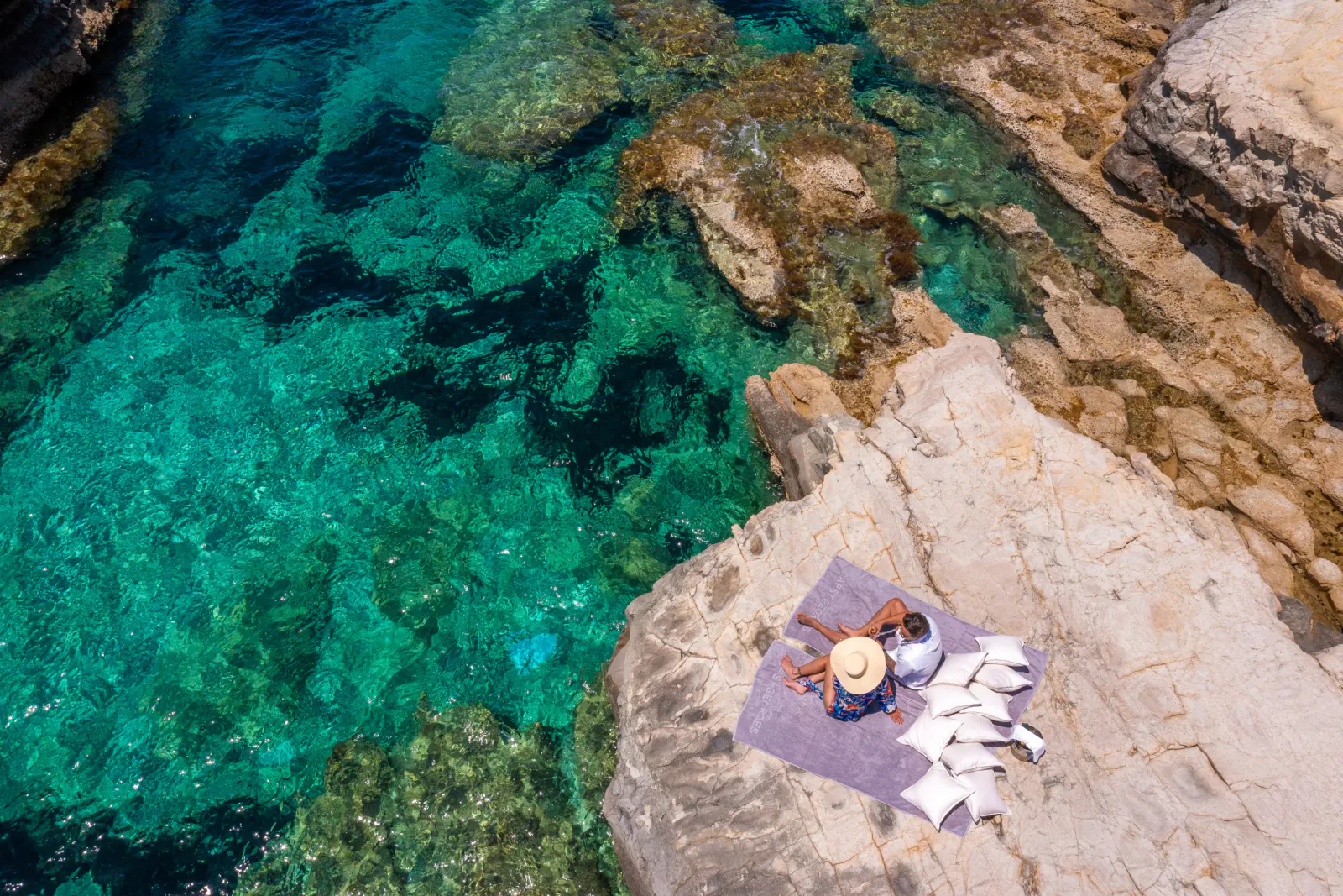 Aerial view of a woman in a straw hat sitting on a towel on a rocky cliff overlooking clear turquoise water at Cap St Georges, Paphos, Cyprus