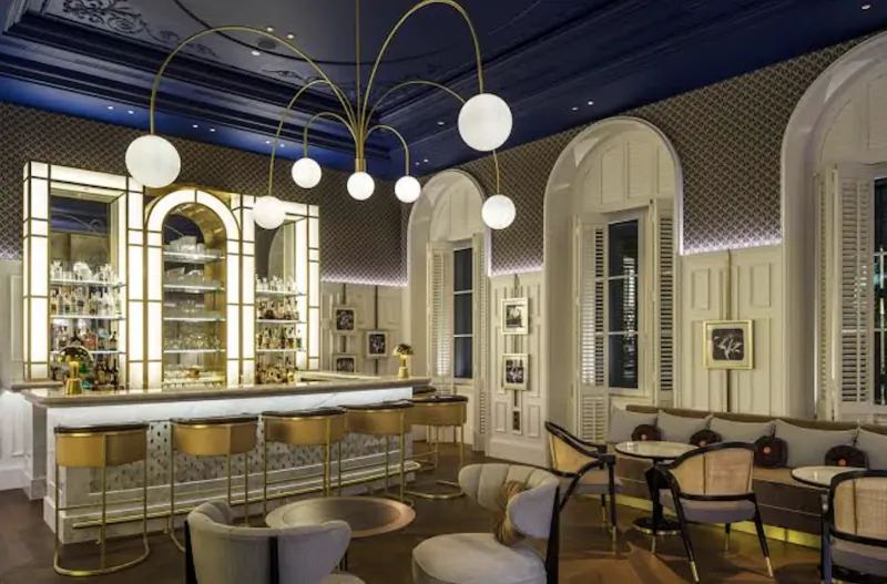 Elegant hotel bar with gold bar stools, arched backlit shelves, and modern chandelier in a blue-ceilinged lounge