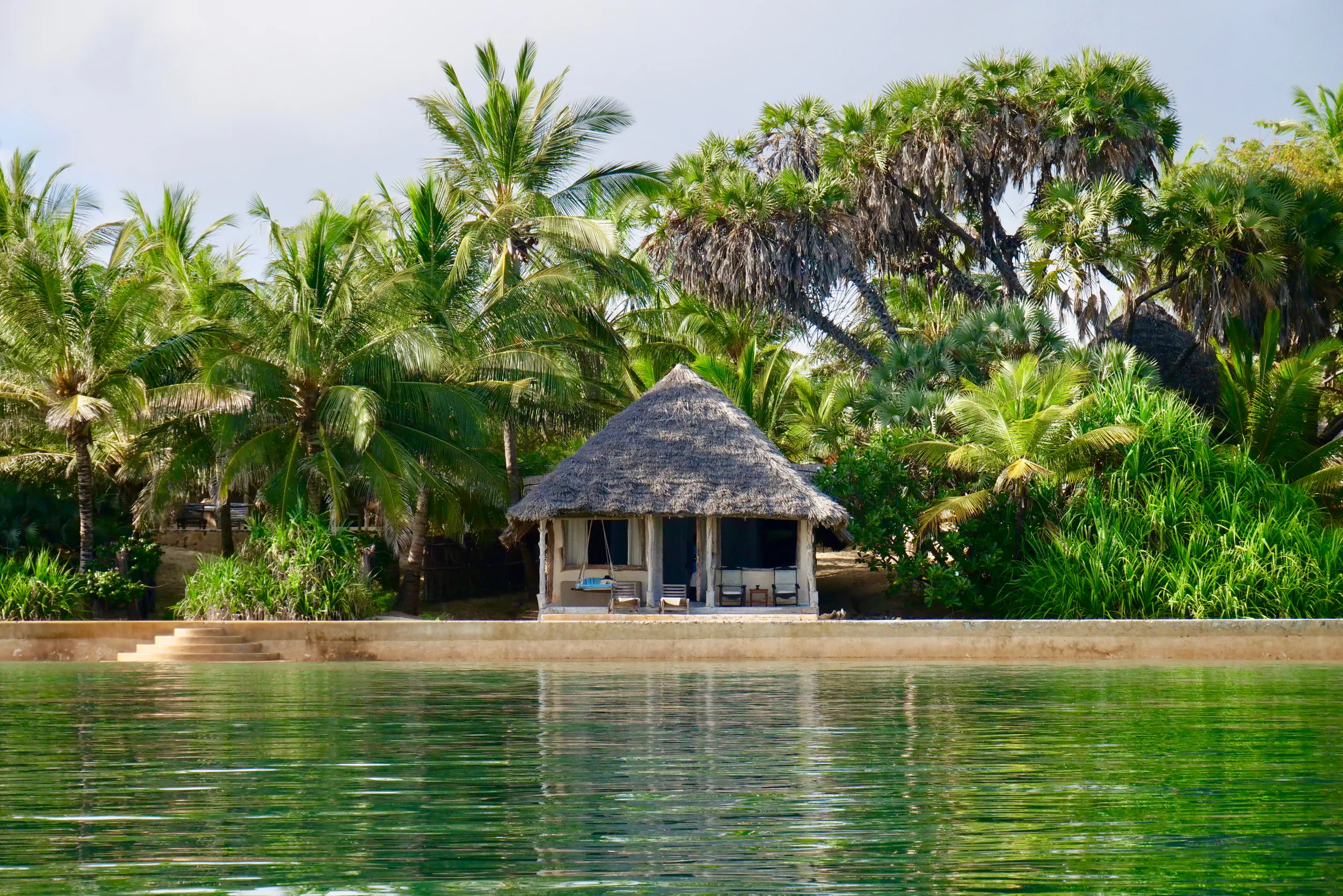 Thatched-roof beach hut with lounge chairs overlooking calm green water, surrounded by lush palm trees at Manda Bay Lodge, Kenya