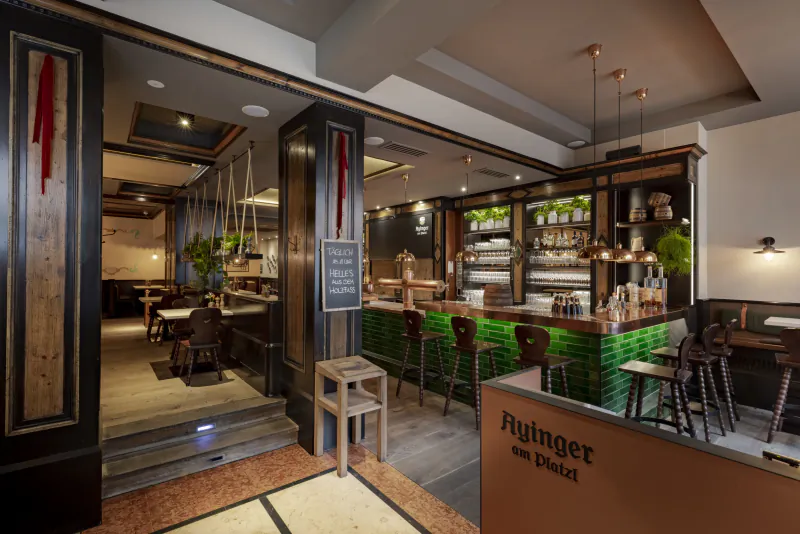 Modern bar interior with green tiled counter, wooden stools, hanging pendant lights, and a sign reading 'Aying am Platzl'