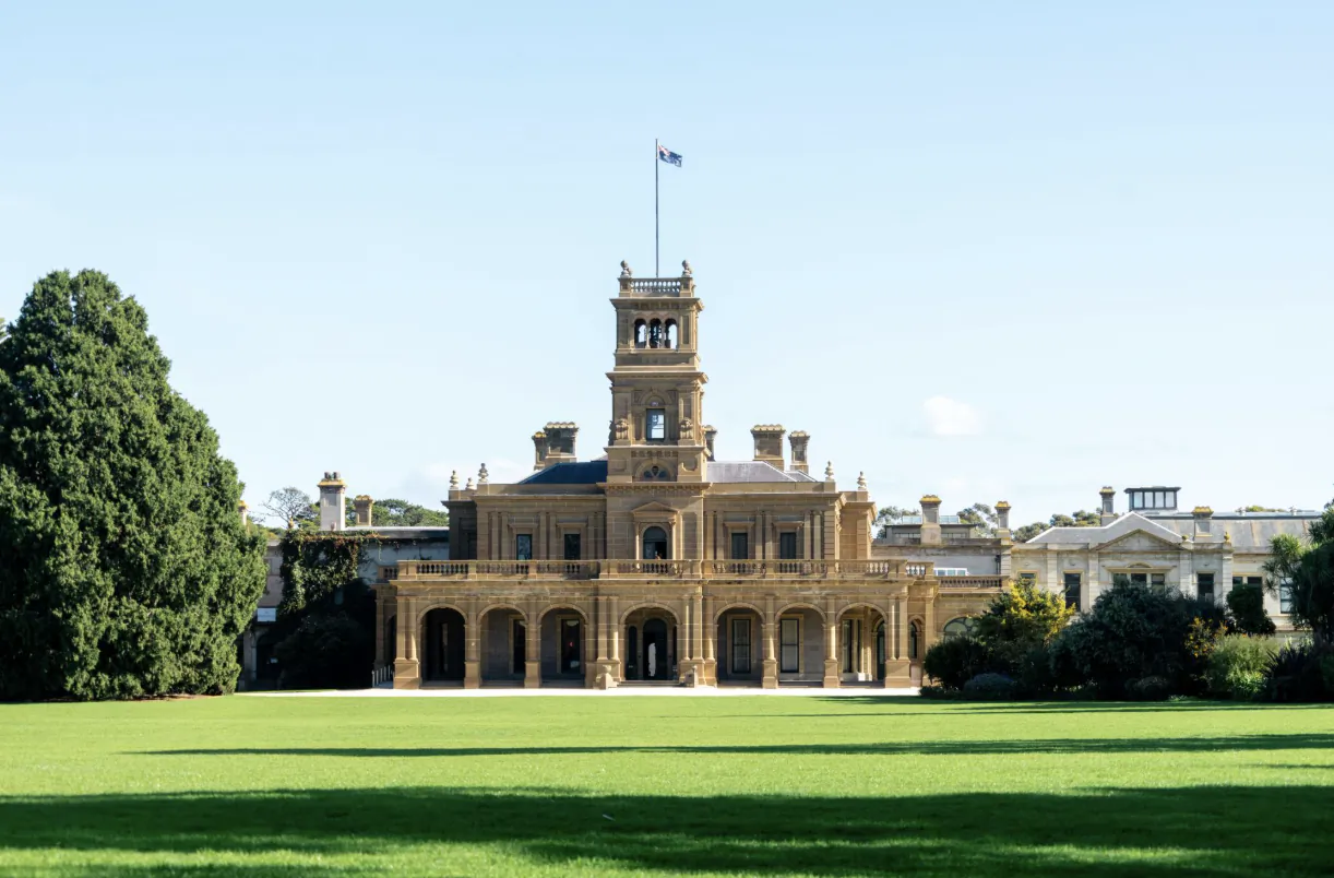 Historic sandstone mansion with central clock tower and Australian flag on manicured green lawn