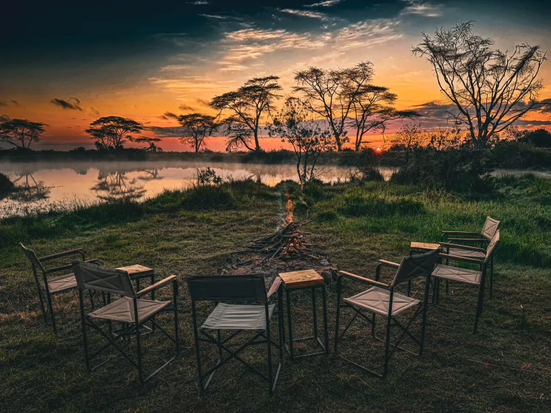Campfire surrounded by empty folding chairs at sunset by a misty lake with acacia trees