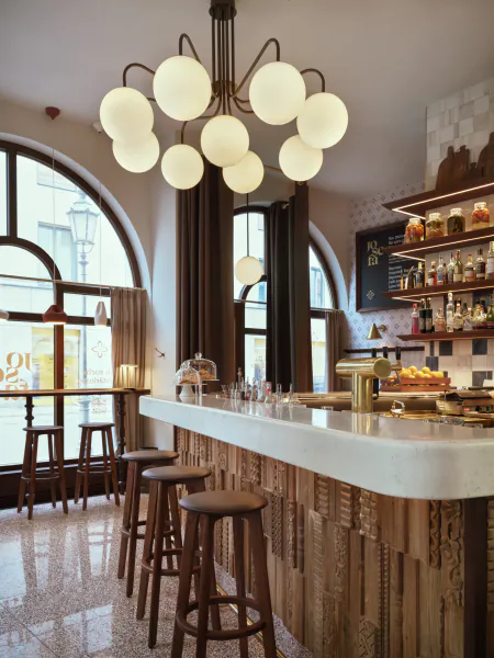 Modern hotel bar with white marble counter, wooden stools, globe chandelier, arched windows, and shelves of bottles and pastries