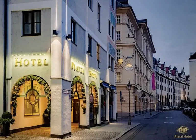 Platzl Hotel Munich facade at dusk with illuminated 'HOTEL' and 'PLATZL HOTEL' signs, arched entrances with floral decorations on a quiet street