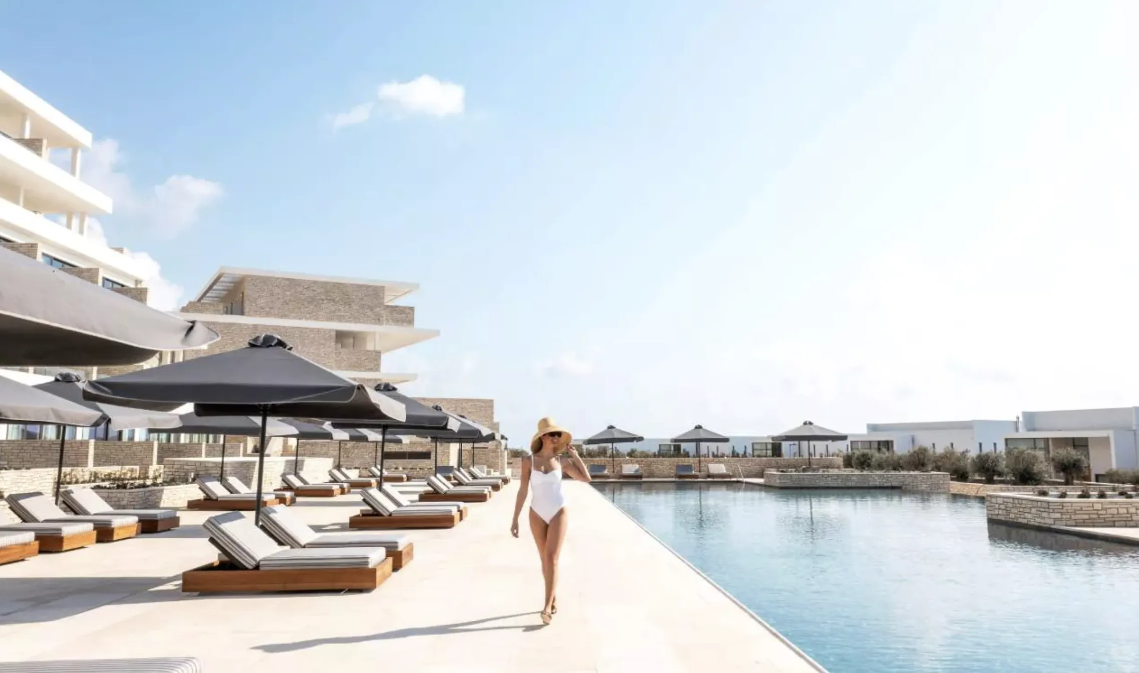 Woman in white swimsuit and hat walking by poolside lounge chairs and umbrellas at luxury resort