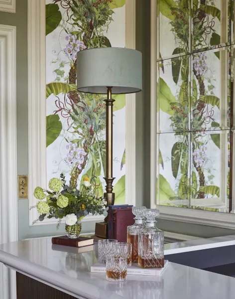 Elegant bar with brass lamp, crystal decanters, floral arrangement, and botanical wallpaper at Hedsor House
