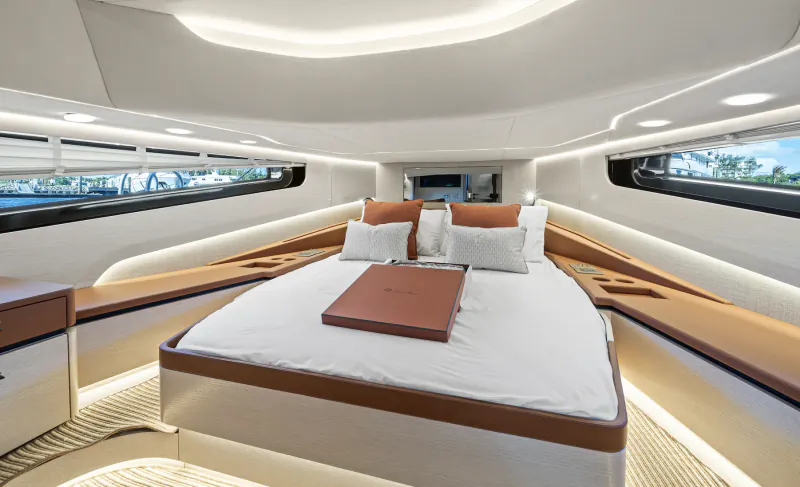 Modern yacht bedroom with large bed, white linens, brown pillows, and a brown tray on the bed, surrounded by curved wood and large windows