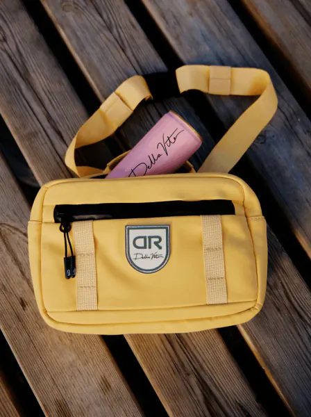Yellow Delta Roam crossbody bag with Della Vite pink bottle on wooden surface