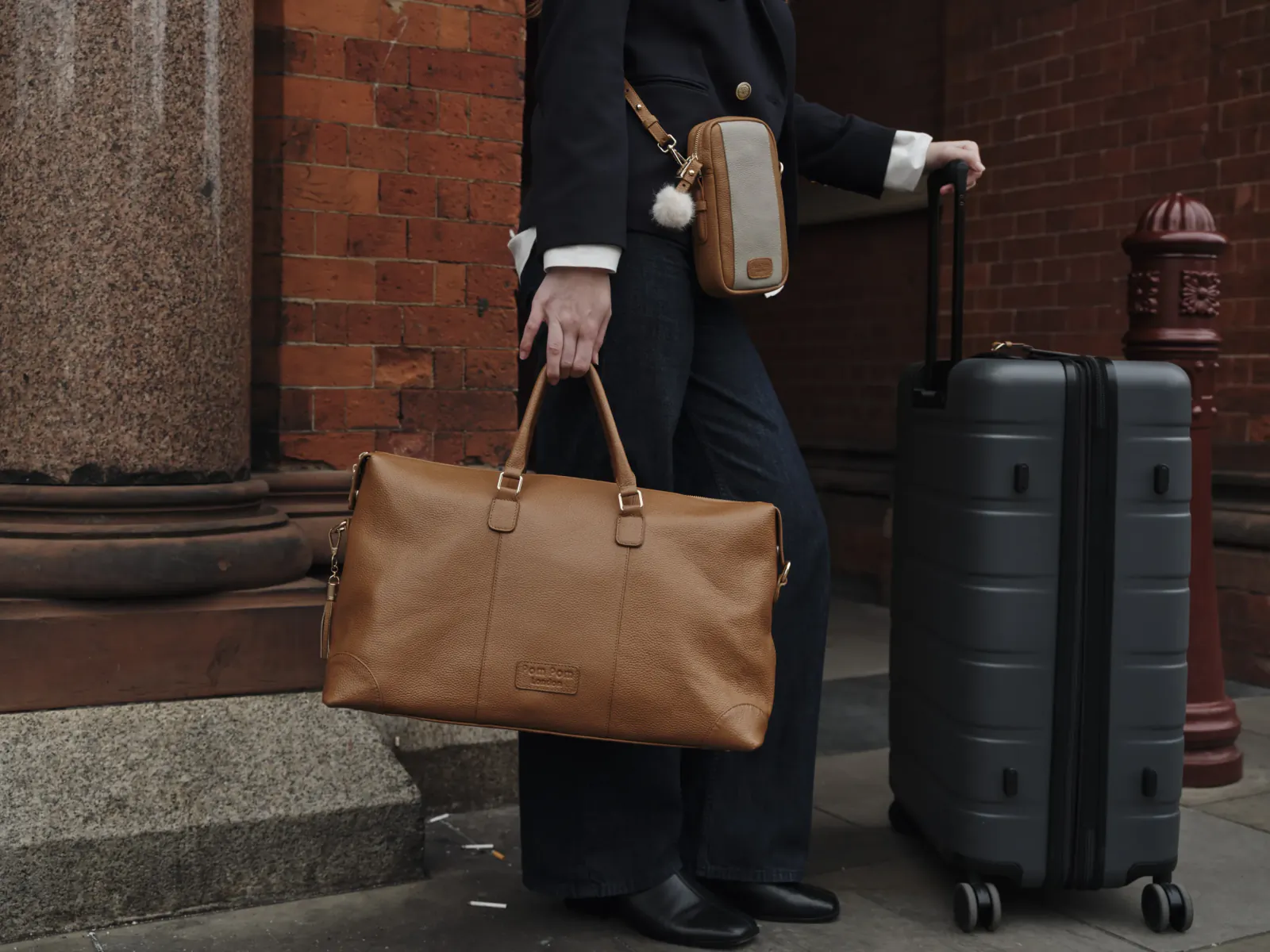 Person in a black coat holding a brown leather duffel bag and rolling a grey suitcase on a city street