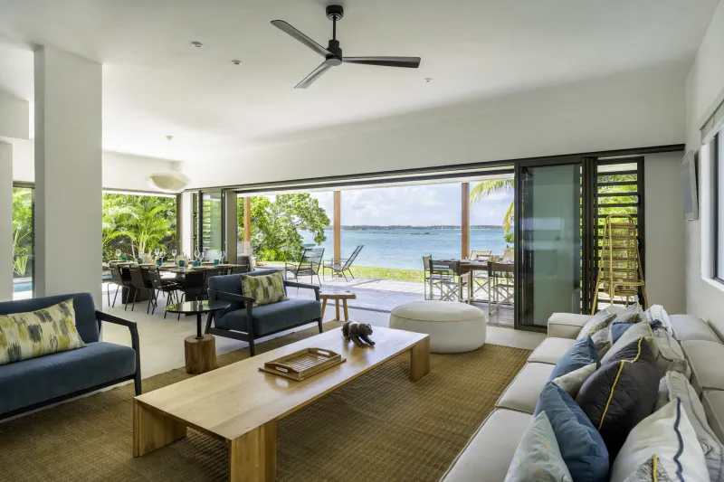 Bright open-plan living room with blue sofas, wooden coffee table, ceiling fan, and large glass doors opening to ocean view and outdoor dining