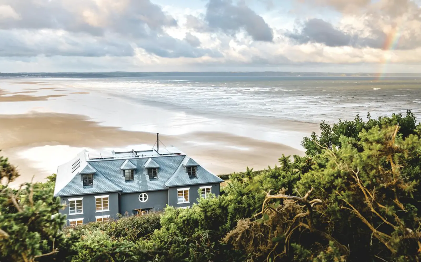Blue coastal house overlooking sandy beach and ocean with rainbow in the sky