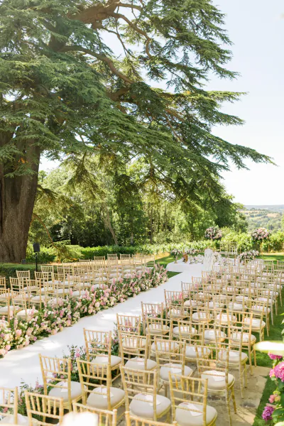 Elegant outdoor wedding ceremony setup with rows of gold chairs and pink floral arrangements under a large tree at Hedsor House