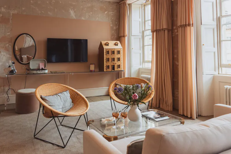 Modern living room with two rattan chairs, glass coffee table with flowers, TV on peach wall, and large windows with peach curtains