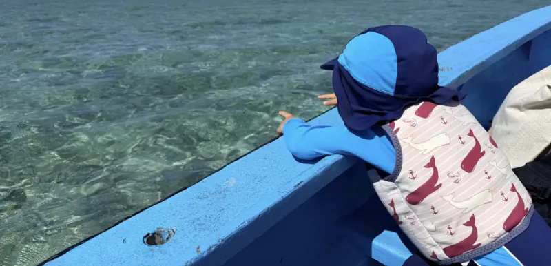Child in sun hat and life vest leaning over blue boat railing, looking at clear turquoise water