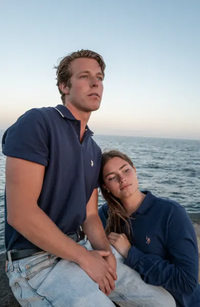 Young man and woman in navy U.S. Polo Assn. polo shirts sitting together by the ocean at sunset