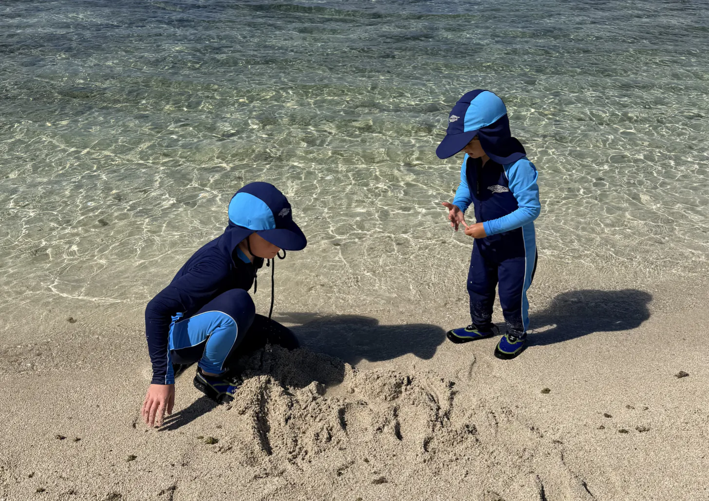 Two young children in blue sun-protective clothing and caps play with sand at the beach edge, one crouching and the other standing in shallow water