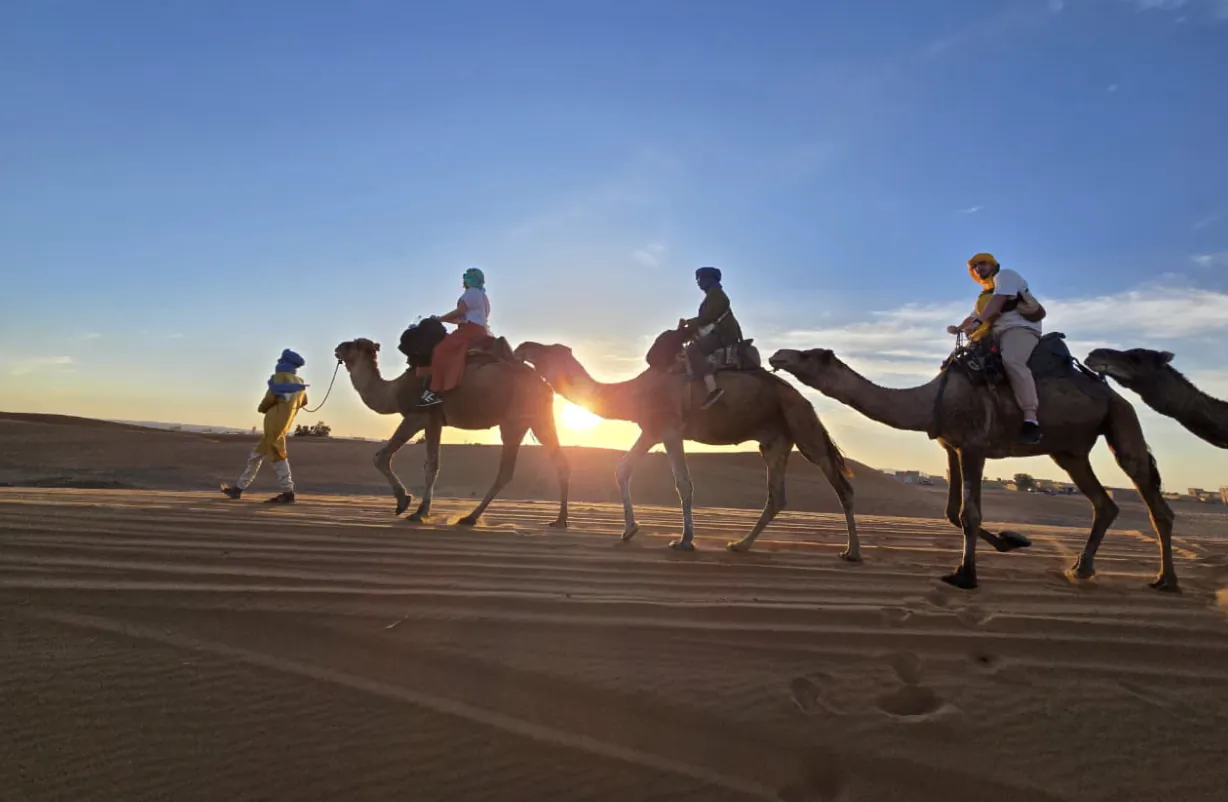Three tourists ride camels in a line across desert dunes at sunset, led by a guide in traditional attire