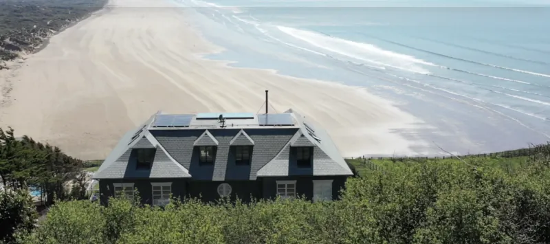 Aerial view of a dark grey house with solar panels overlooking a wide sandy beach and ocean waves