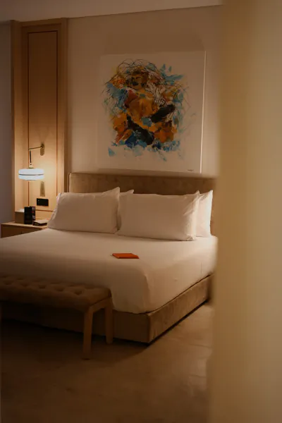 Modern hotel bedroom with large bed, white linens, abstract orange-blue painting above headboard, and wooden bench at foot