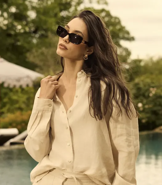 Woman in cream linen shirt and sunglasses posing outdoors by a pool
